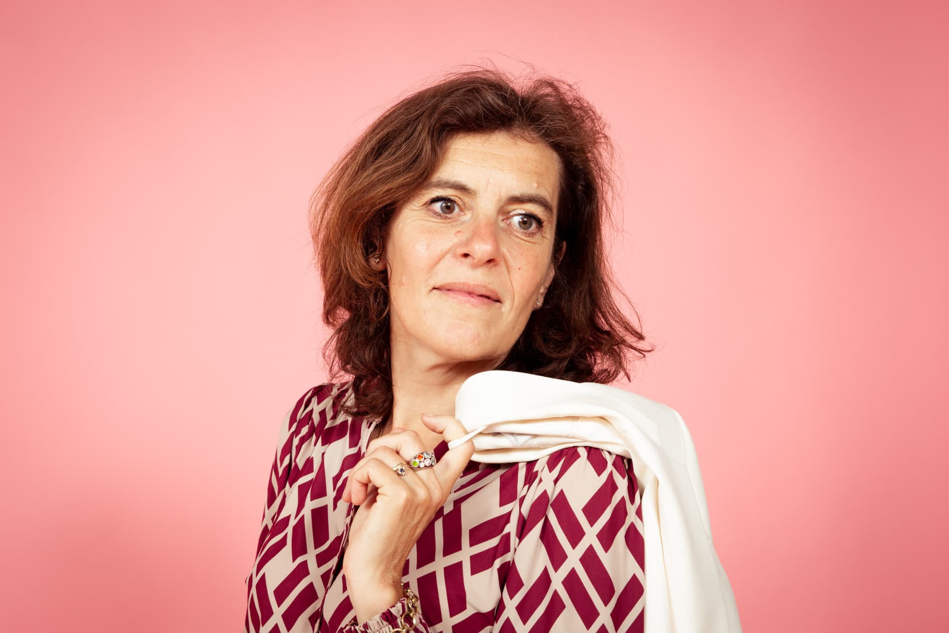 Emily Staphorst kijkt in de camera met haar colbert over haar schouder, tegen een roze achtergrond